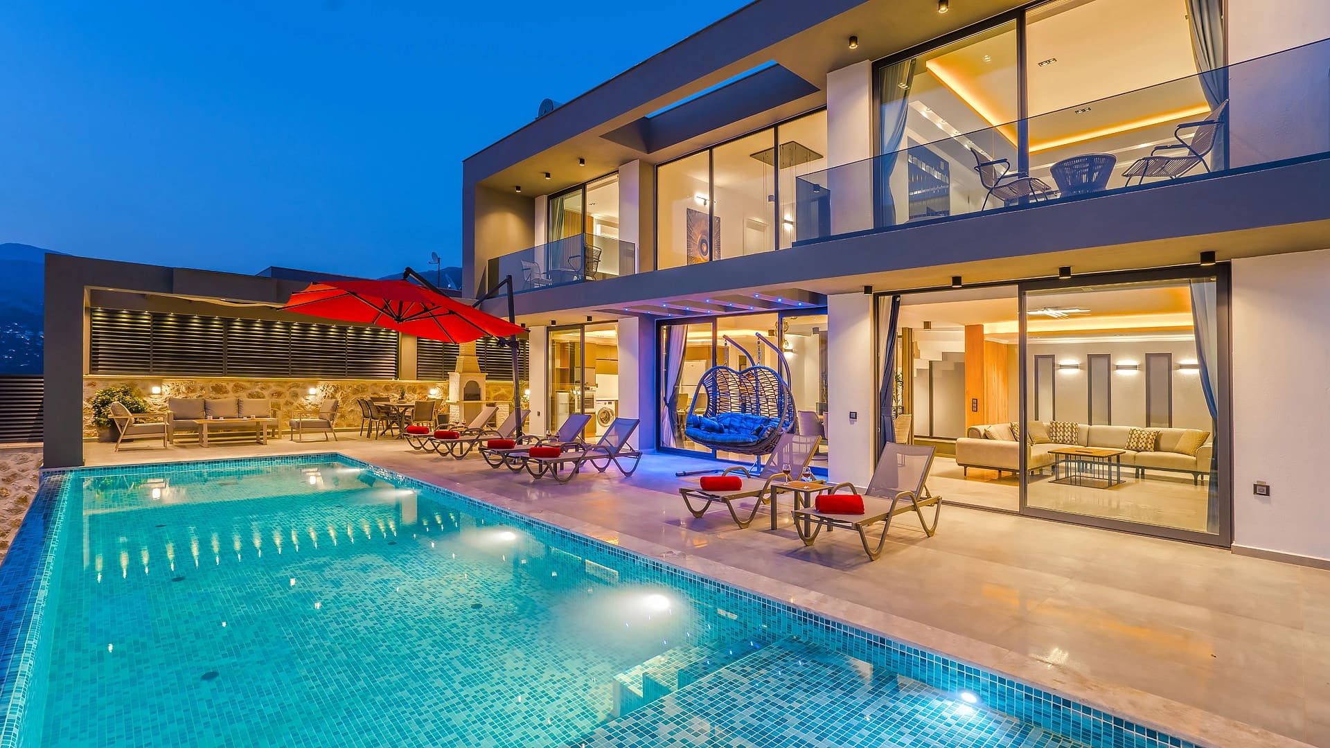 Akşam ışıklandırmalı iki katlı modern villa, önünde geniş yüzme havuzu ve şezlonglar.