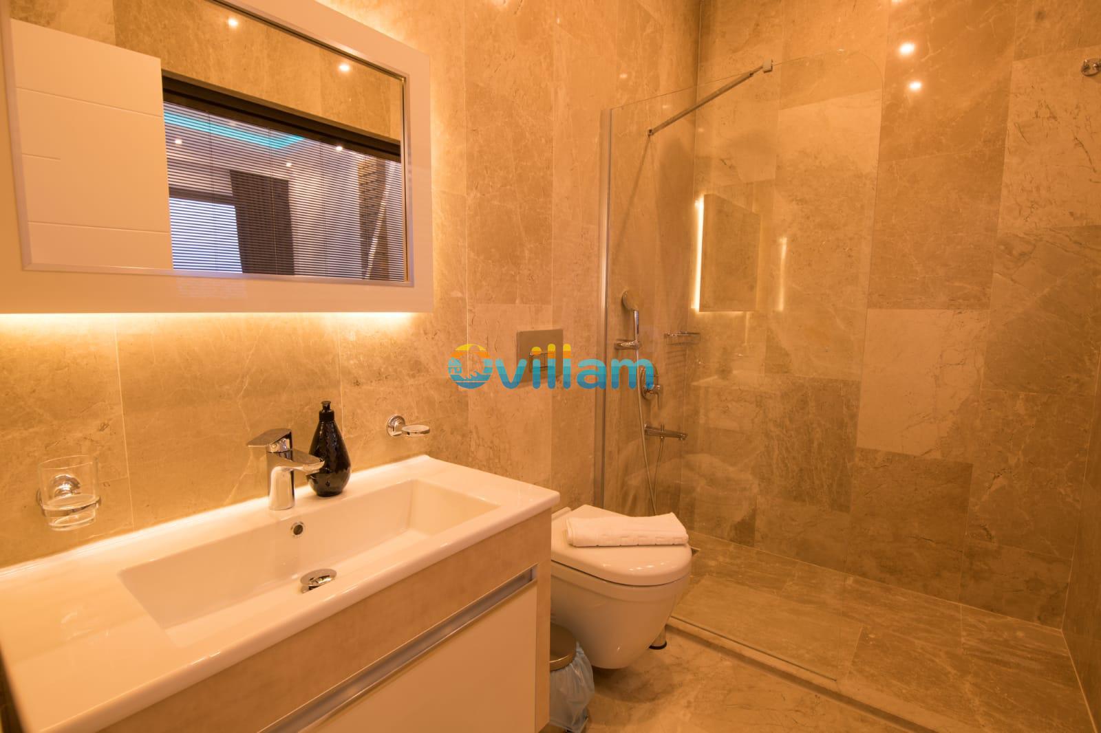 Modern banyo içerisinde beyaz lavabo, ayna ve cam duşakabin.