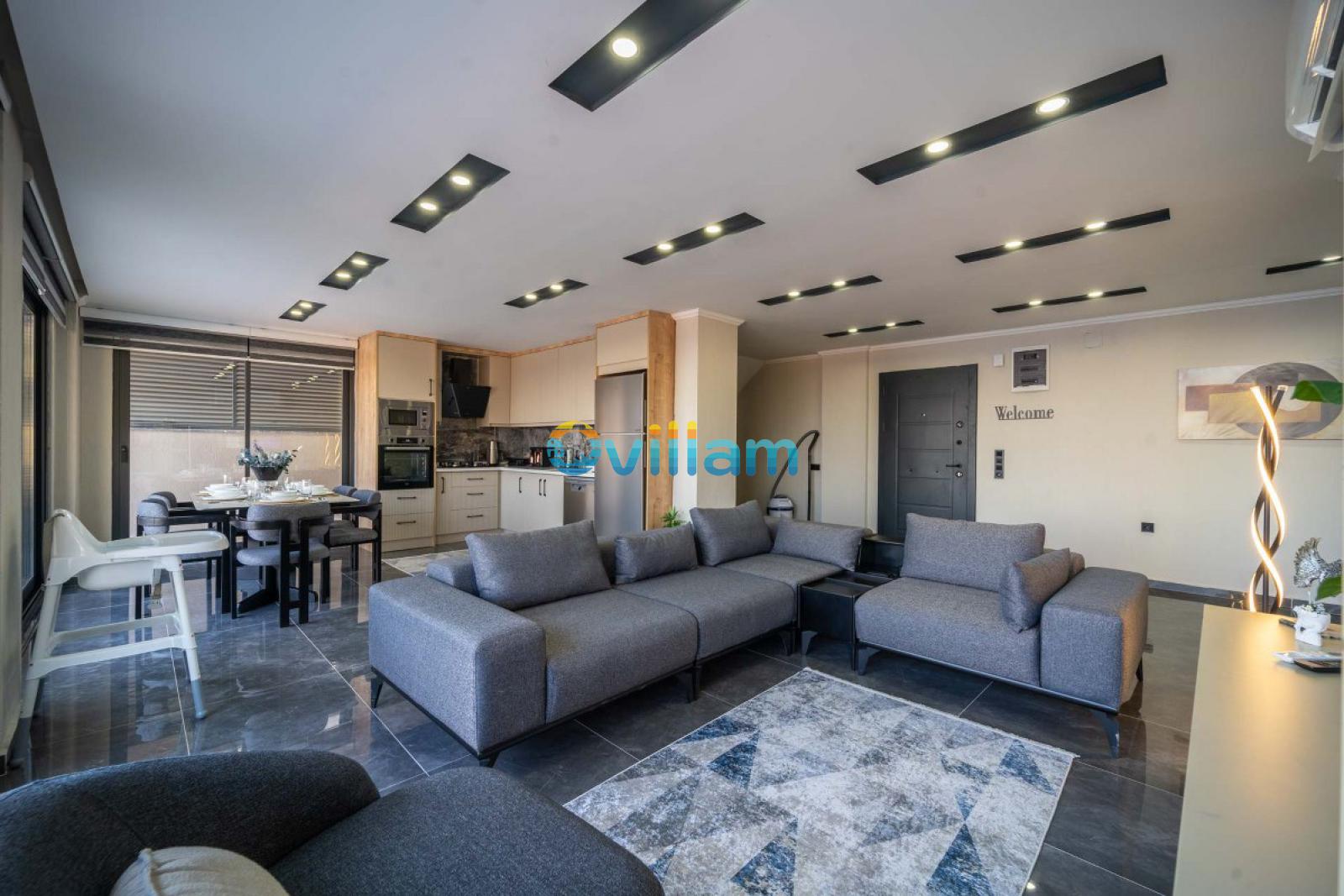 Modern mutfaklı açık plan salon, gri köşe koltuk ve şık tavan aydınlatmaları.