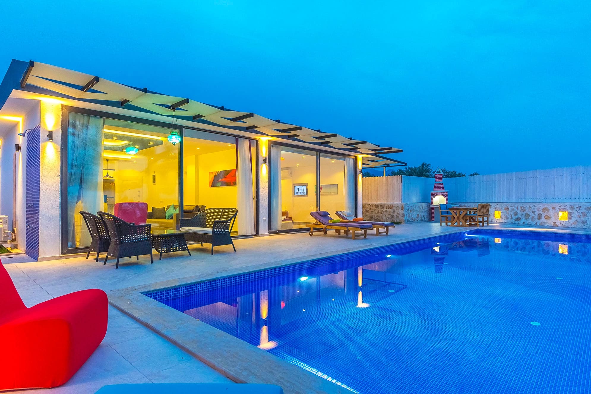 Akşam saatlerinde ışıklandırılmış yüzme havuzu, veranda ve villanın cam cephesi.