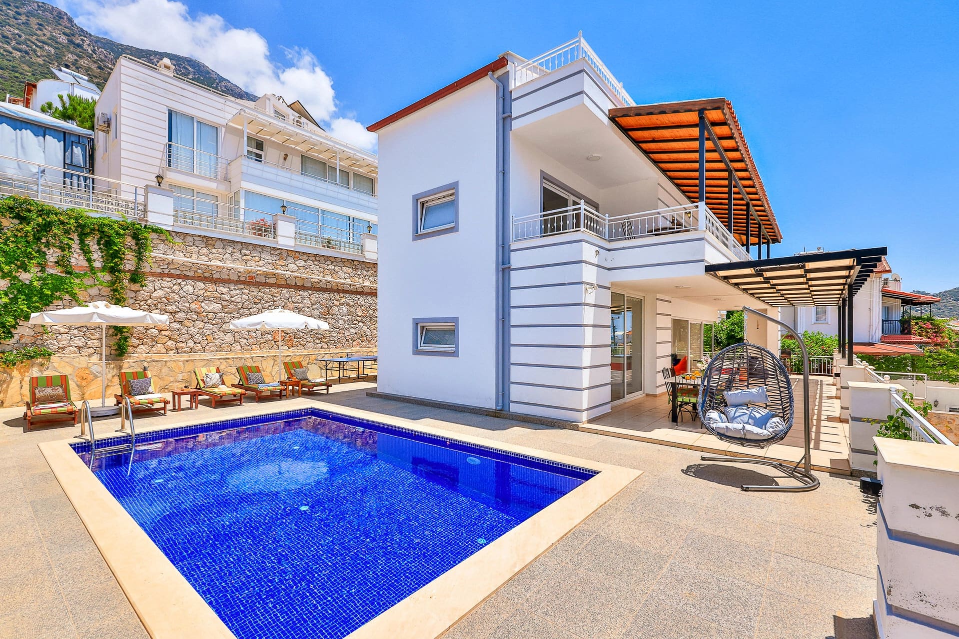 Mavi mozaikli havuzu, şezlongları ve salıncağı olan iki katlı beyaz villa.