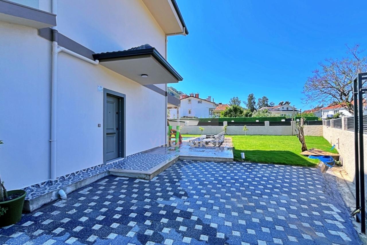 Desenli taş döşeli ön avlu, gri giriş kapısı ve arka tarafta çim alan bulunan villa dış cephesi.