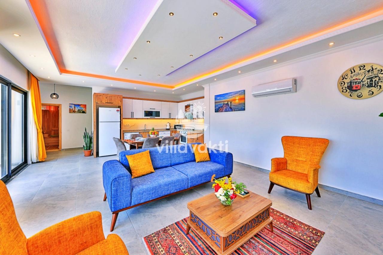 Mavi koltuk, turuncu berjerler ve ahşap sehpanın yer aldığı açık planlı modern salon ve mutfak.
