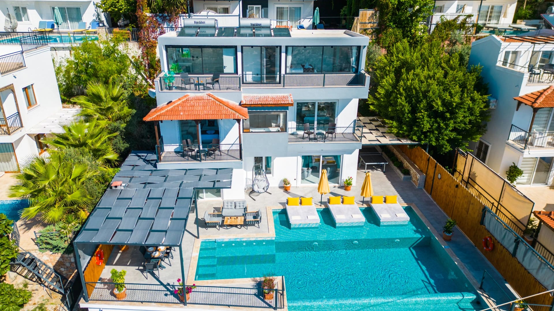 Üst açıdan geniş yüzme havuzlu, sarı şemsiyeli ve çardaklı üç katlı modern beyaz villa.