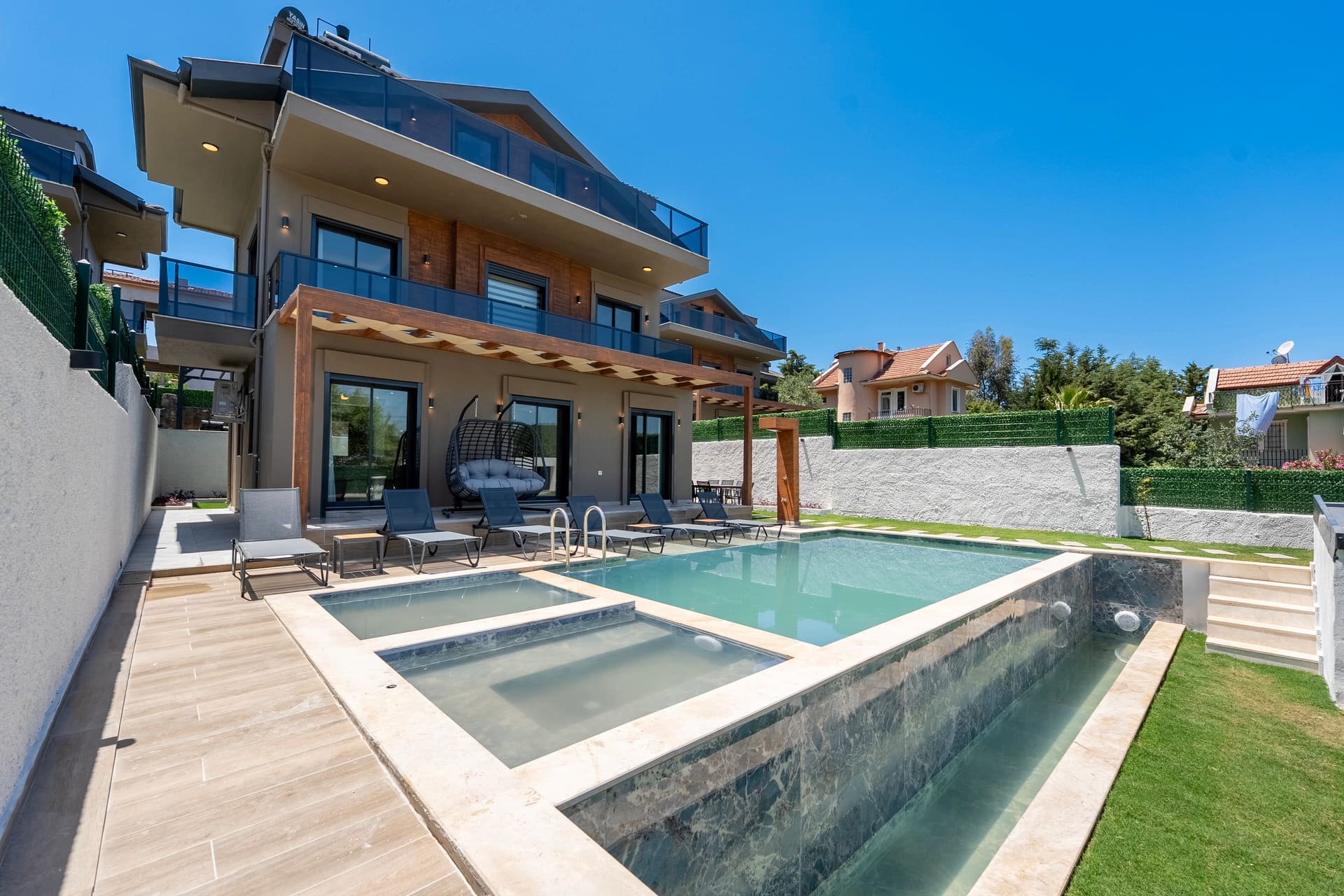 Açık mavi yüzme havuzu, şezlonglar ve çim alanı bulunan çok katlı modern villa.