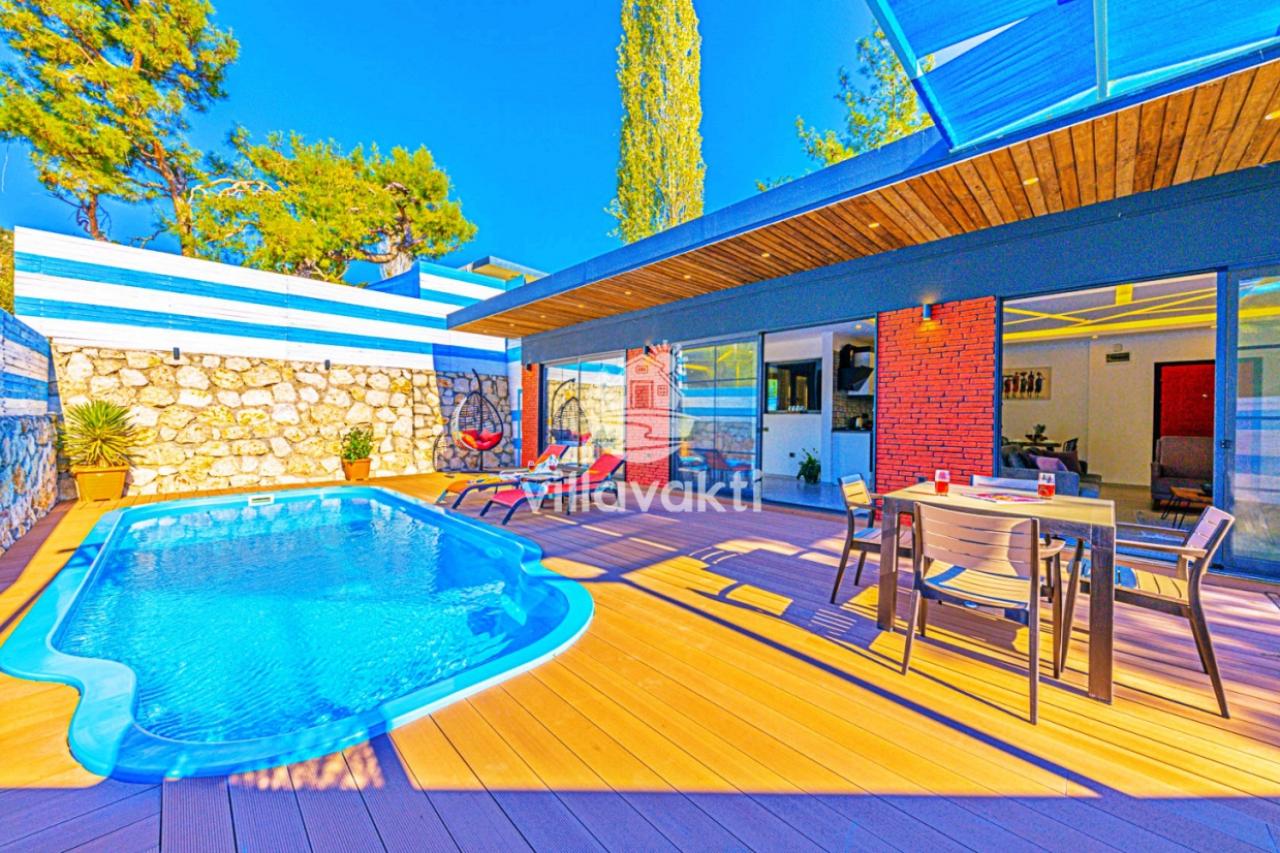 Ahşap zeminli verandada özel yüzme havuzu, yemek masası takımı ve modern villa cephesi.
