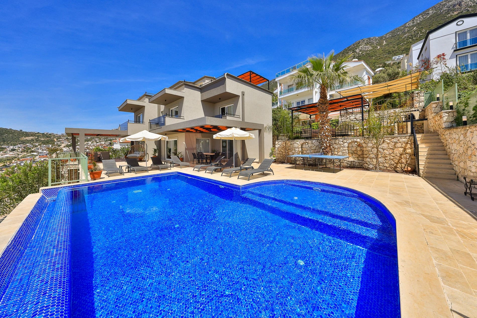 Geniş yüzme havuzu, şezlonglar, masa tenisi ve arkada modern iki katlı villa.