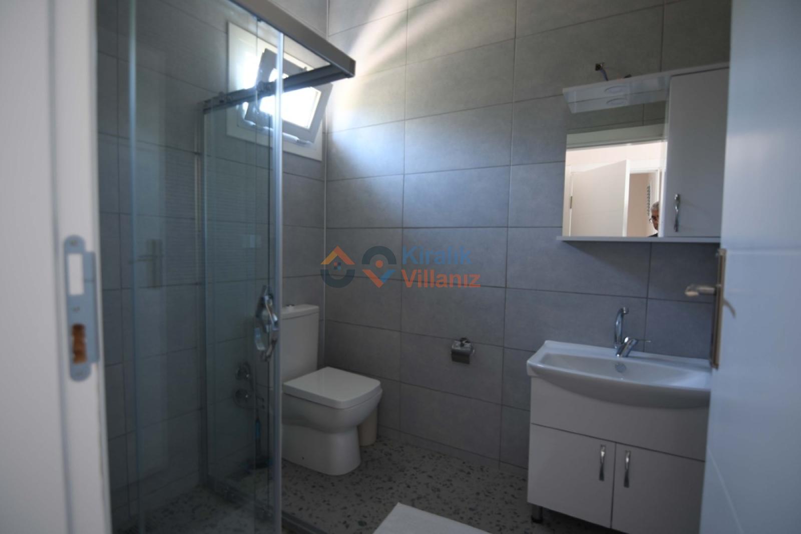 Gri seramik kaplı banyoda cam kabinli duş, klozet ve dolaplı lavabo var.