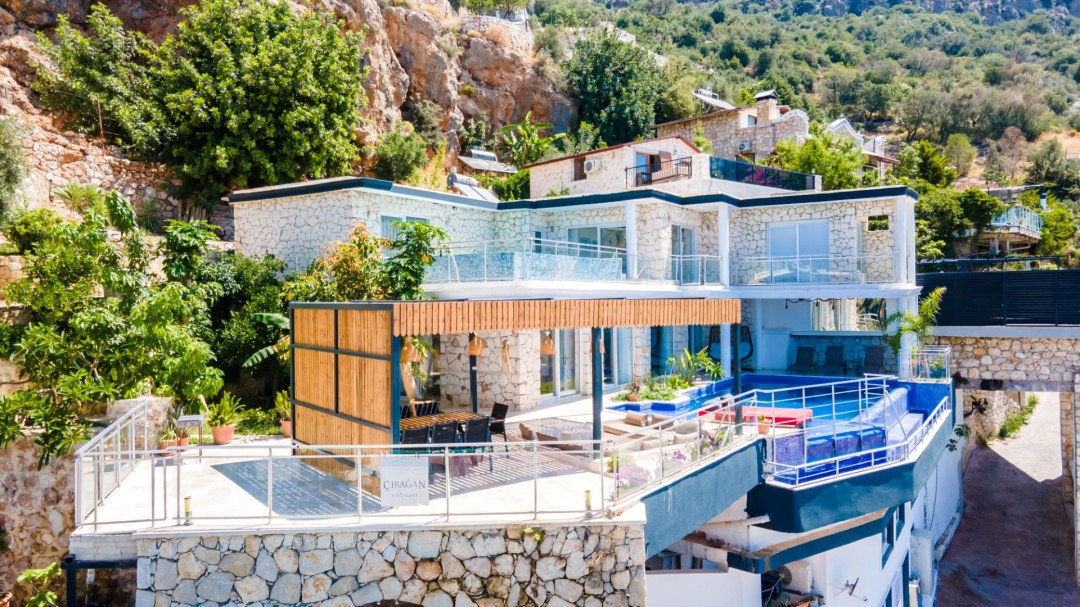 Kayalık yamaç önünde taş duvarlı, havuzlu ve ahşap pergolalı iki katlı villa.
