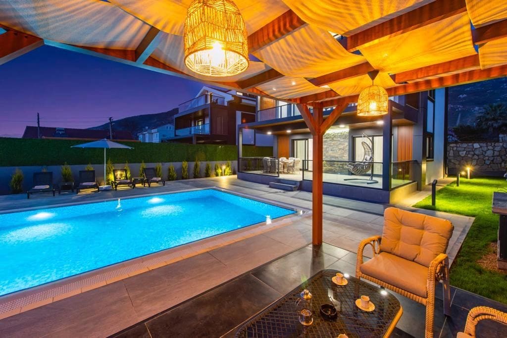 Akşam aydınlatmalı, gölgelikli oturma alanı ve havuzlu villa bahçesi.