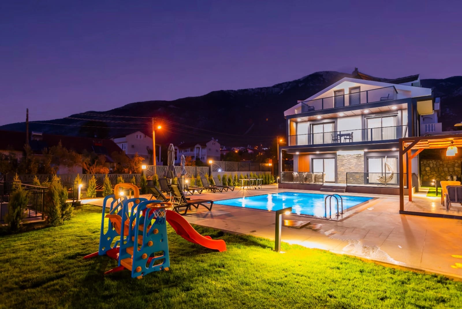 Akşam ışıklandırmalı havuz kenarında çocuk kaydırağı ve arkada villa dış cephesi.