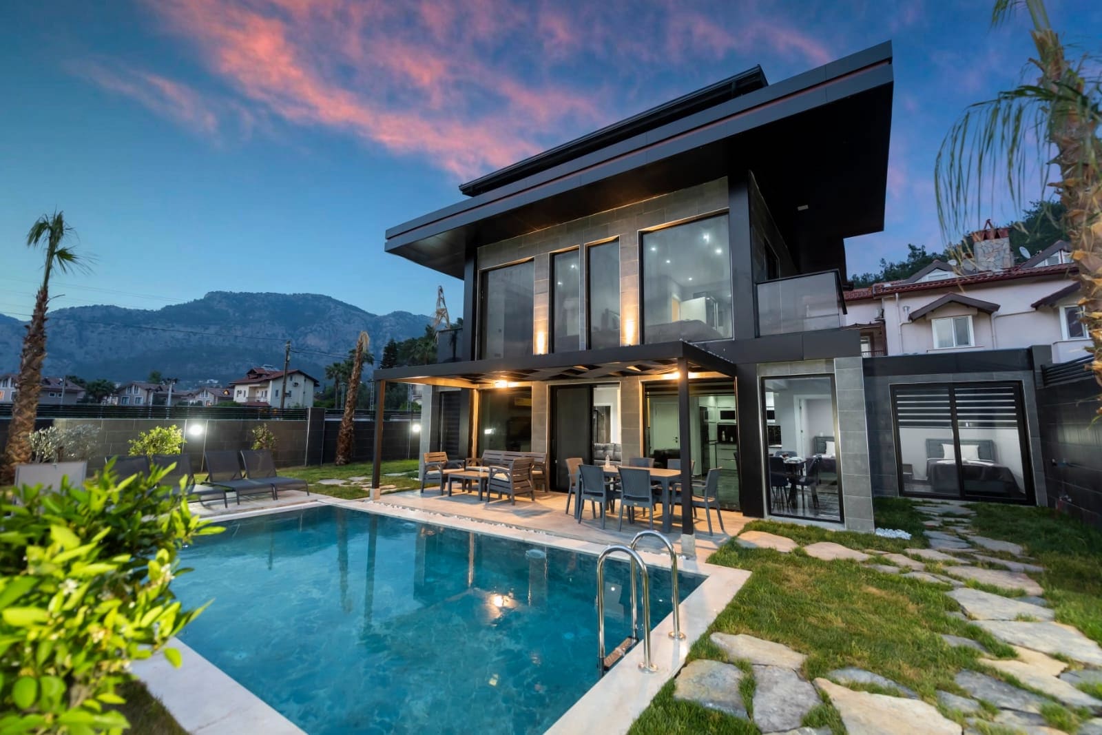 Akşam karanlığında dağ manzaralı havuz ve bahçesi olan modern villa dış cephesi.