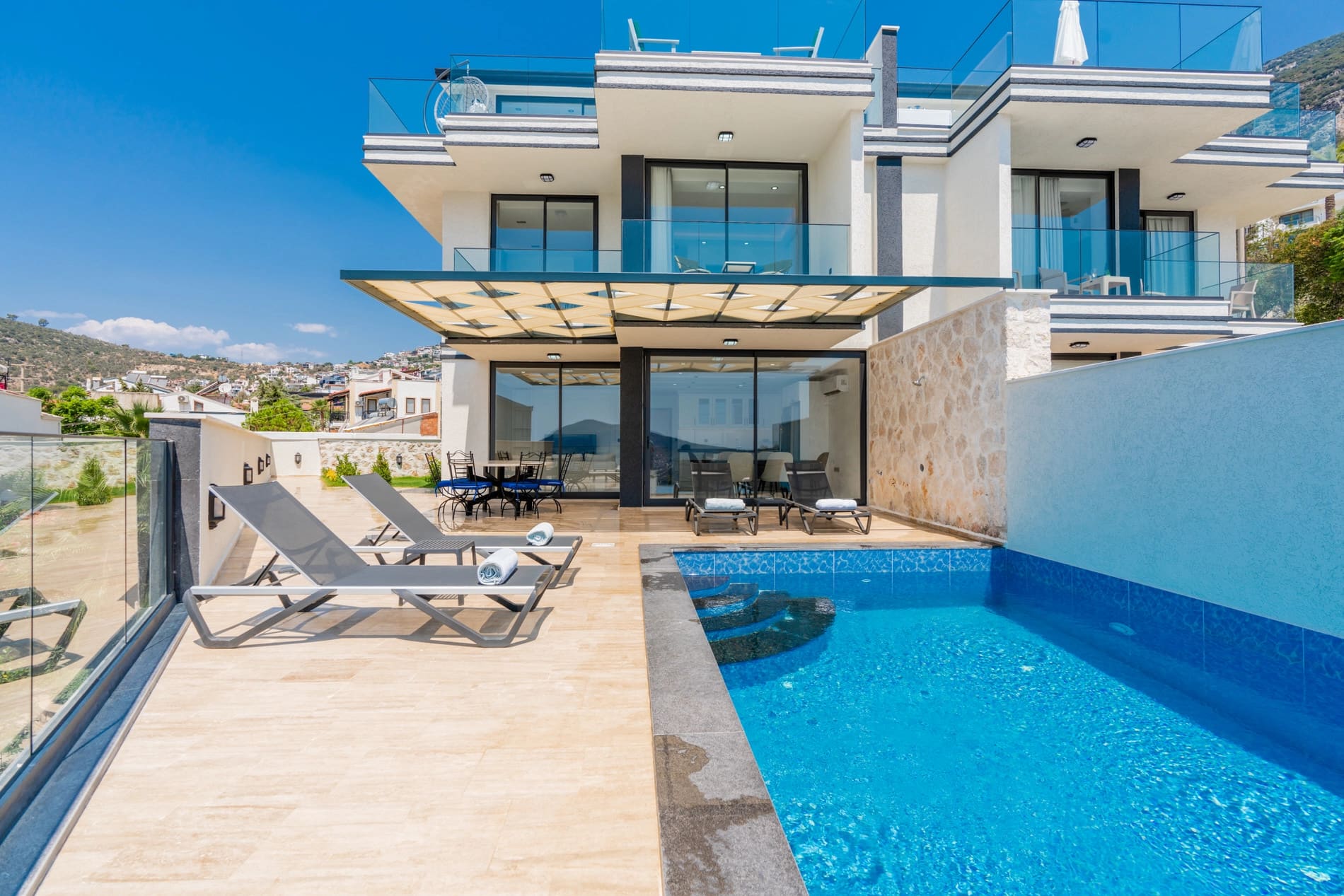 Gündüz saatlerinde havuz başında şezlonglar bulunan modern villa dış cephesi.