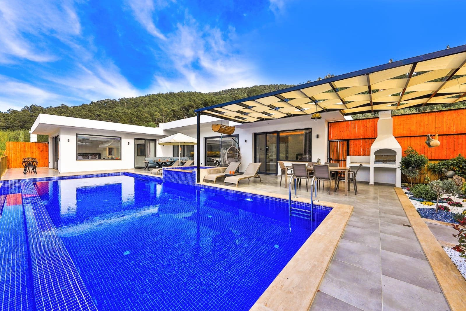 Açık havuz kenarında yemek masası, şezlonglar ve şemsiyeler bulunan modern villa.