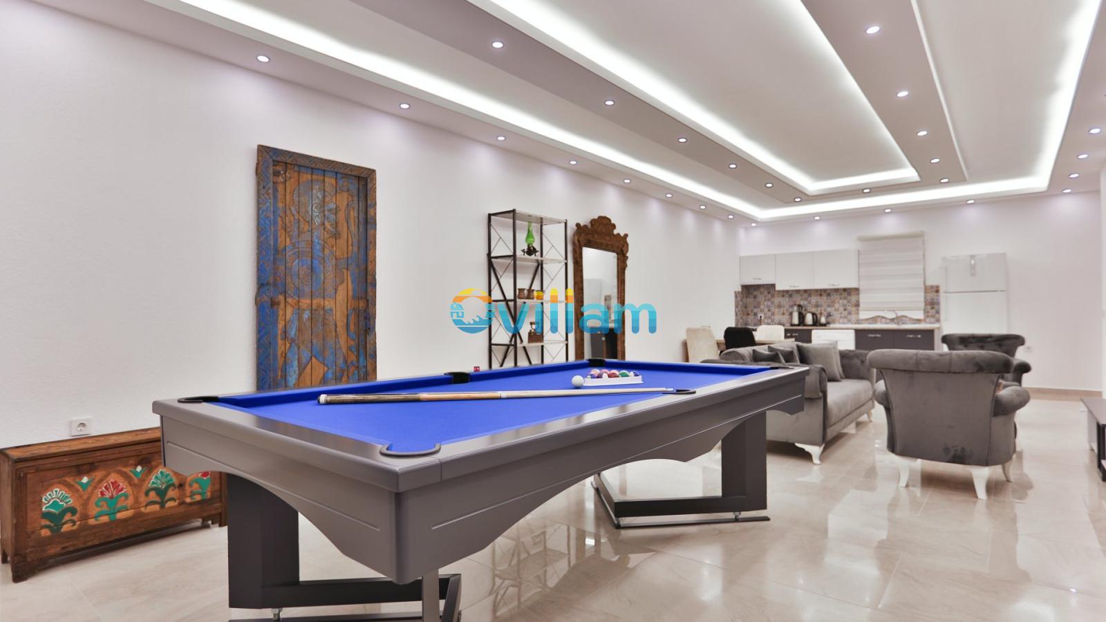 Geniş salonda mavi çuhalı bilardo masası, koltuklar ve modern tavan aydınlatması.