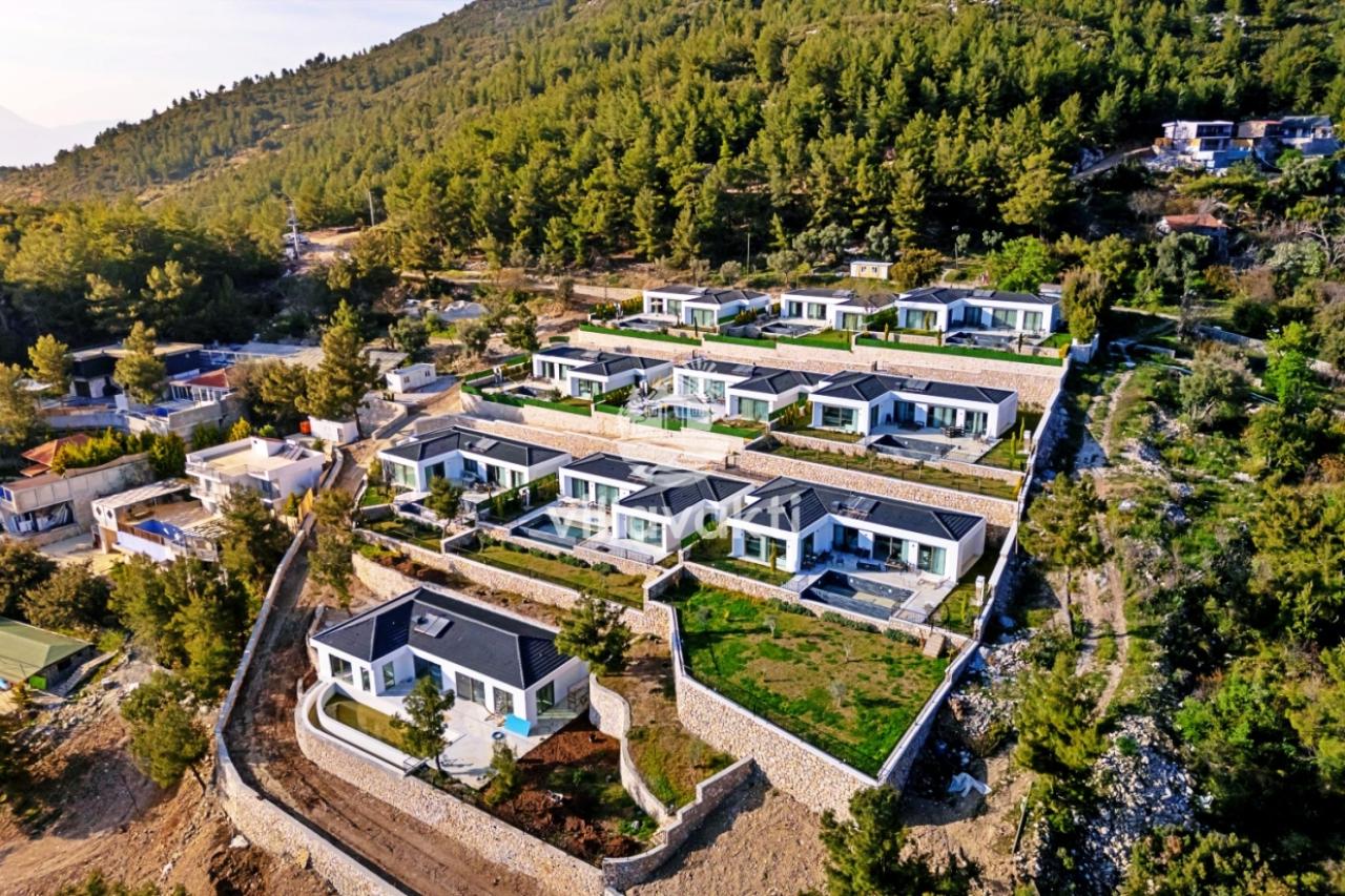 Ormanlık dağ yamacında kademeli olarak yerleşmiş havuzlu villaların havadan görünümü.