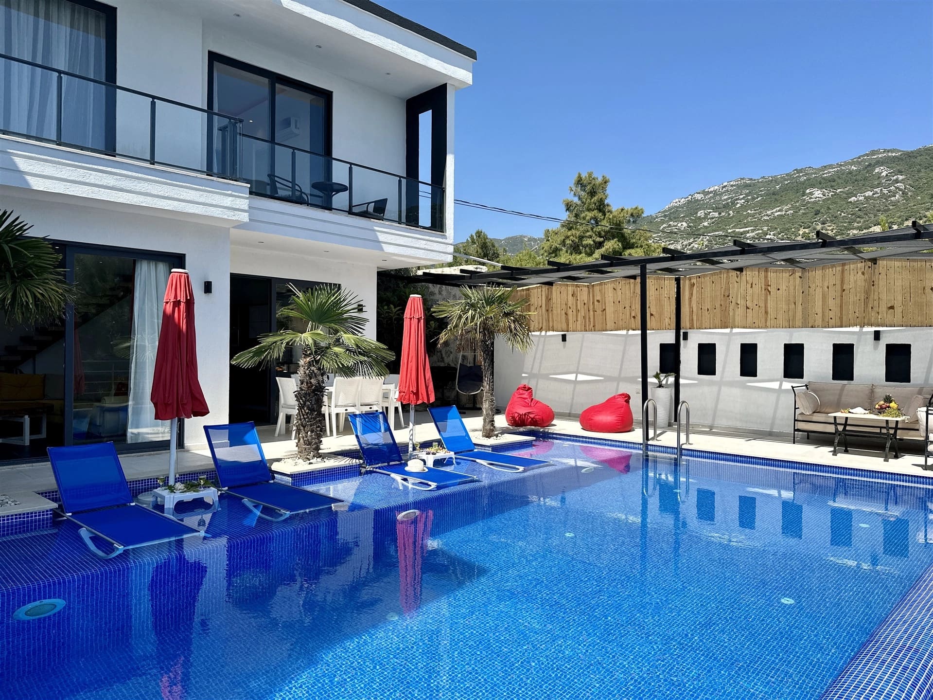 Mavi şezlonglu havuz ve arka planda dağ manzaralı beyaz villa dış cephesi.