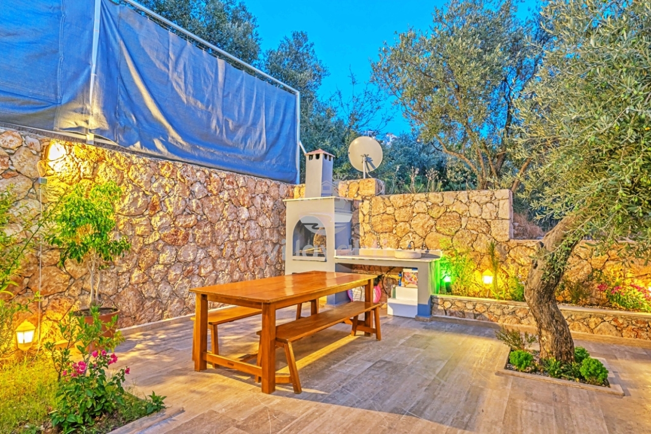 Bahçede ahşap yemek masası, taş duvar ve barbekü alanı.