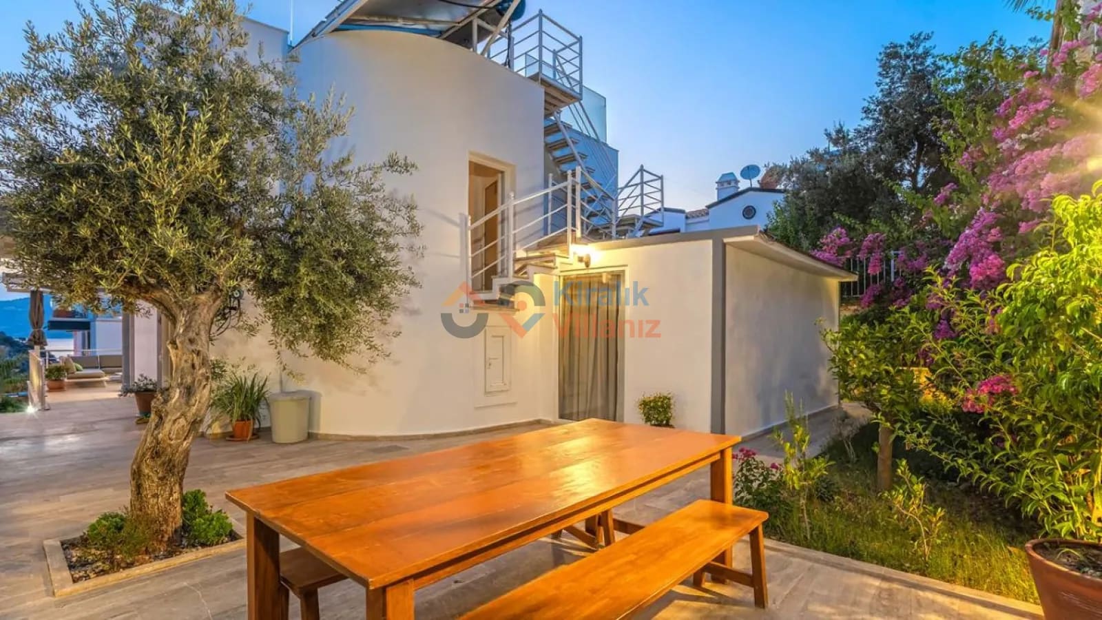 Bahçede ahşap yemek masası ve zeytin ağaçları bulunan villa dış cephesi.