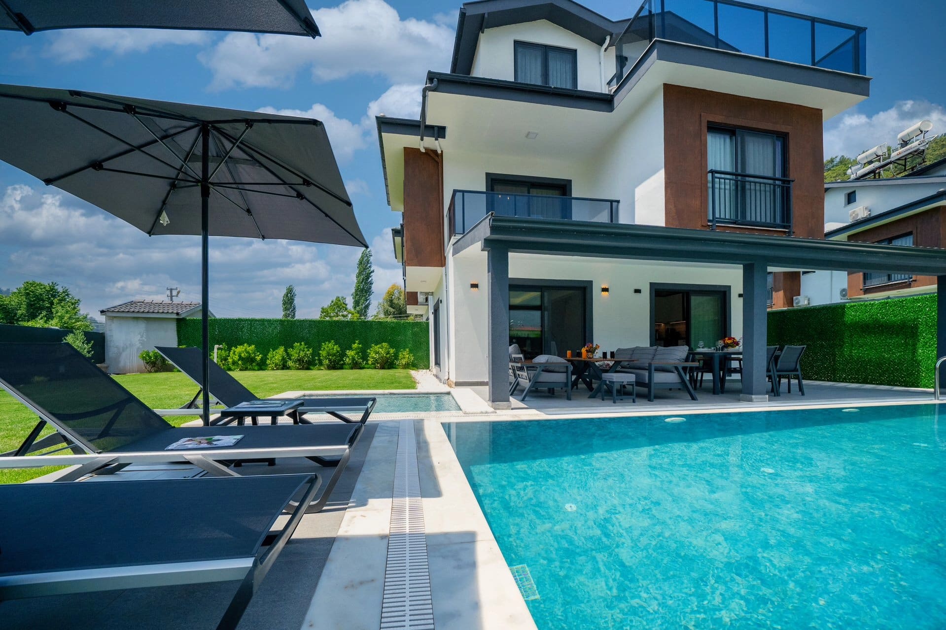 Gündüz saatlerinde havuz kenarında şezlonglar ve şemsiyeler bulunan iki katlı modern villa.