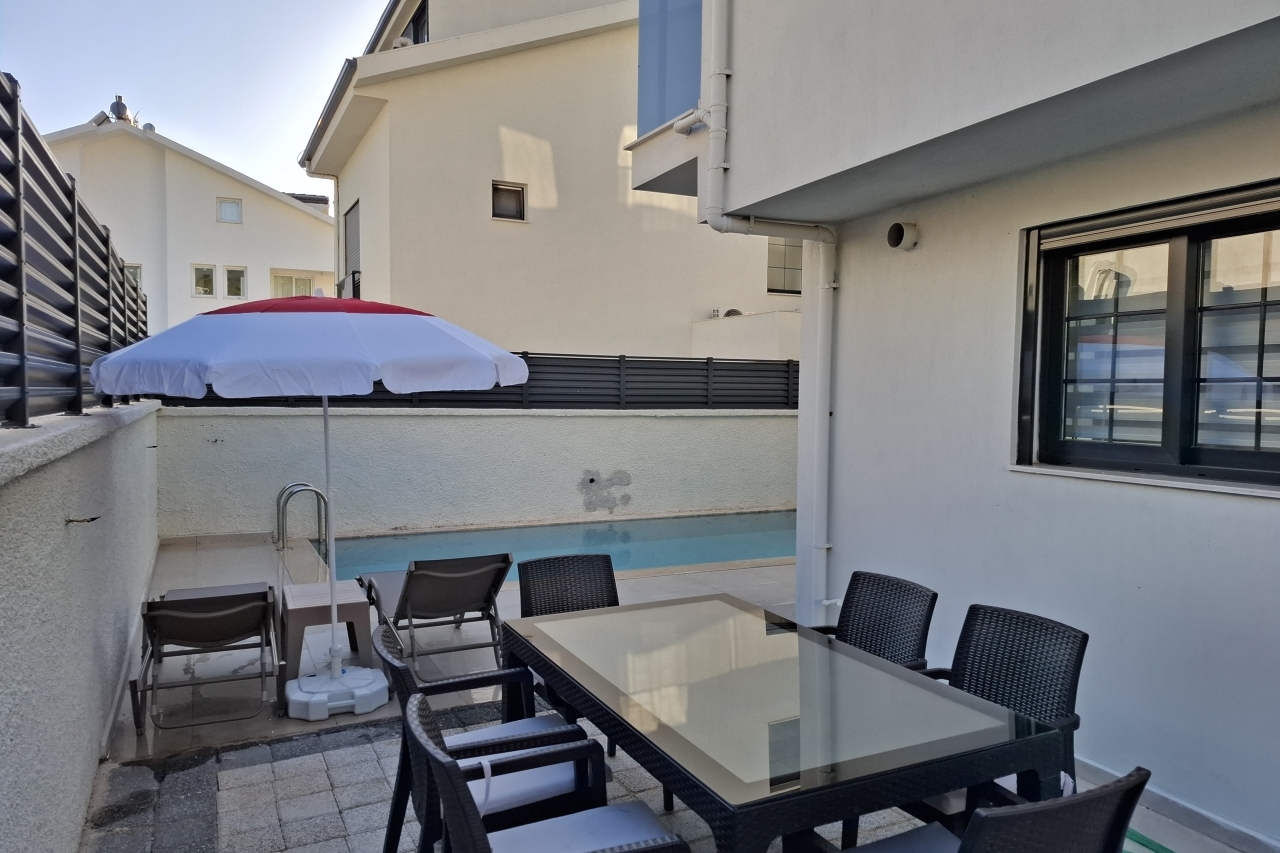 Bahçede yemek masası ve havuz kenarında şezlonglar bulunan villa dış mekanı.