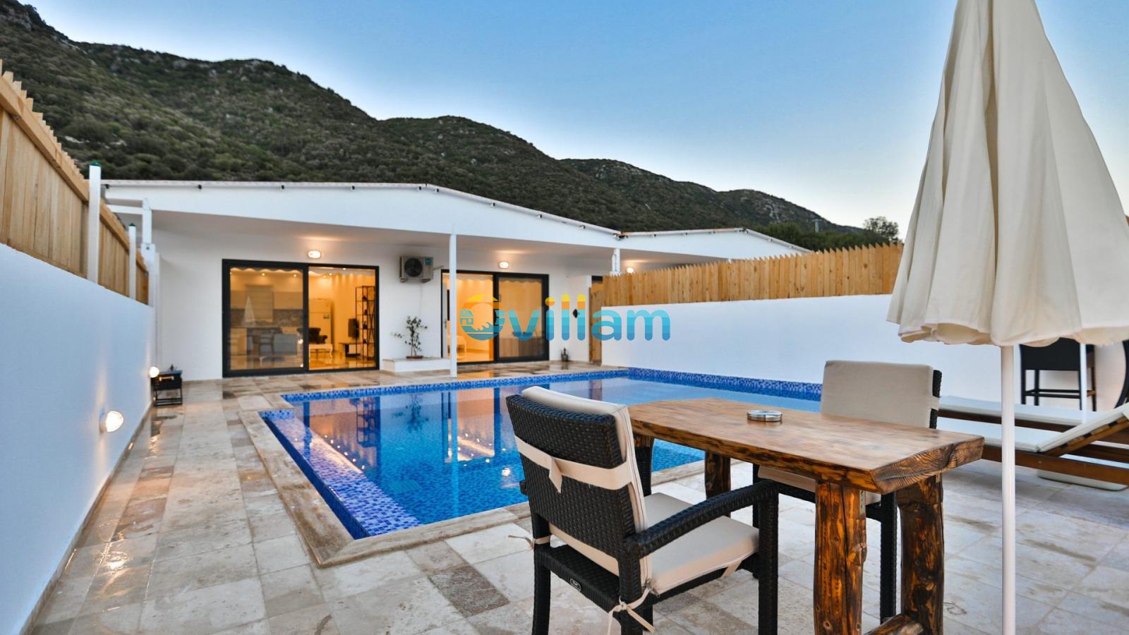 Havuz kenarında masa, sandalyeler ve arka planda dağ manzarası bulunan villa bahçesi.