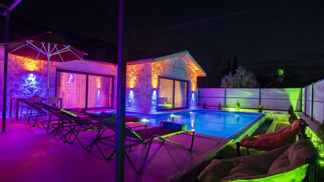 Gece vakti mor ve sarı dış aydınlatmalı taş villanın havuzu ve şezlongları.