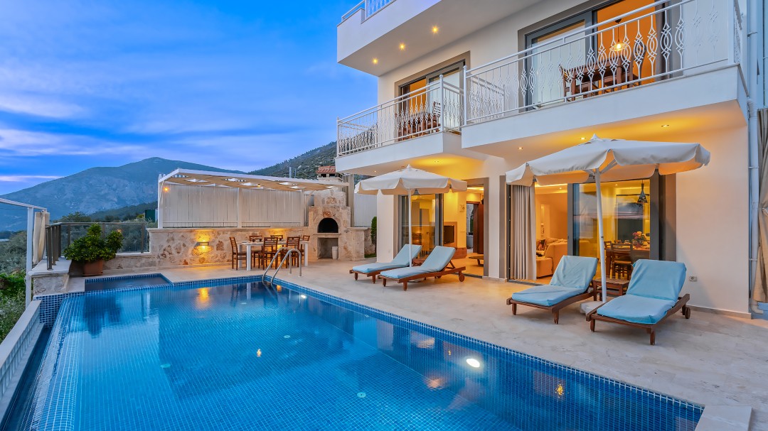 Akşam vakti havuz kenarında şezlonglar, şemsiyeler ve masalar bulunan iki katlı beyaz villa.