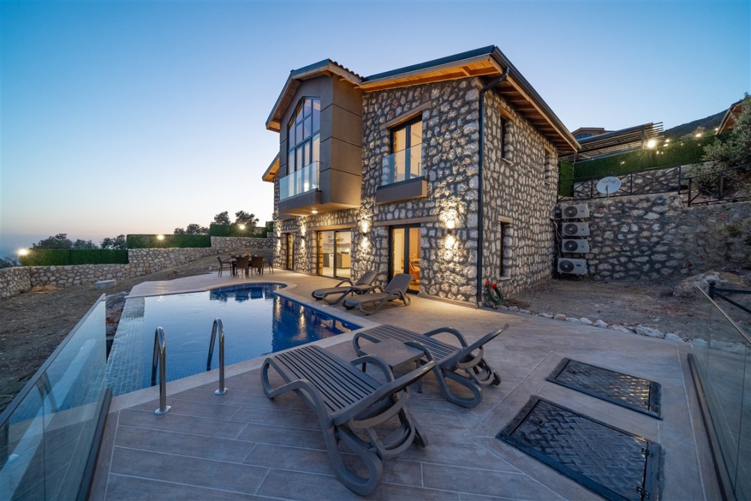 Akşam karanlığında dış cephesi aydınlatılmış taş villa, havuzu ve şezlongları.