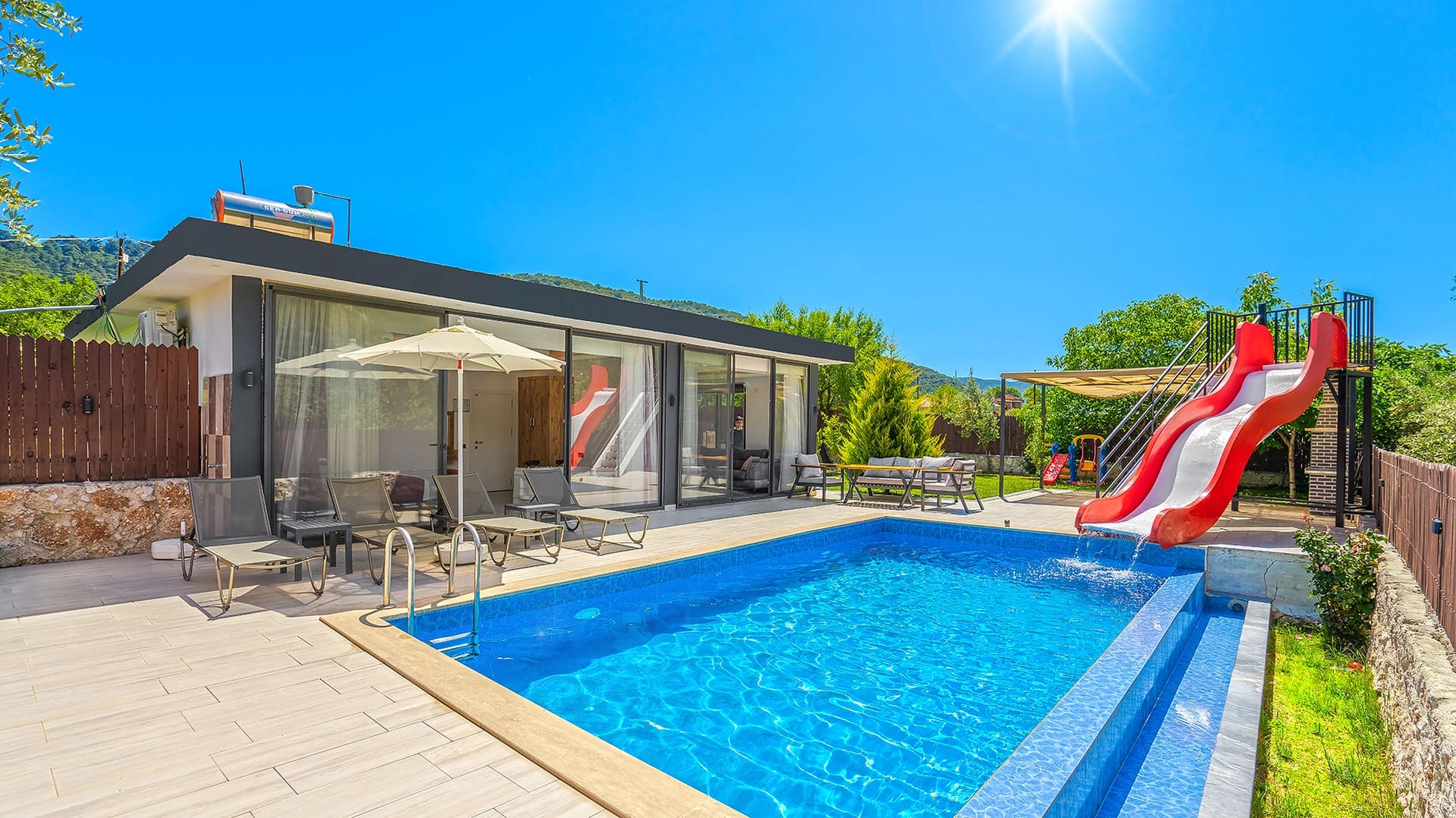 Havuz kenarında kırmızı kaydırak, şezlonglar ve güneş şemsiyesi bulunan tek katlı villa.