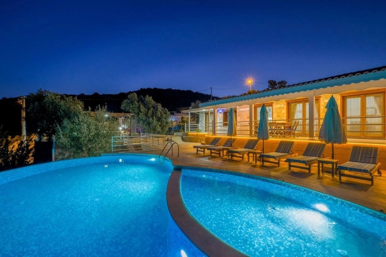 Gece aydınlatmalı havuz kenarında şezlonglar ve taş villa dış cephesi.