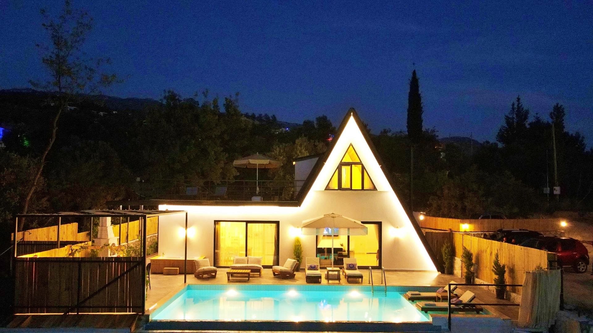 Gece çekiminde aydınlatılmış üçgen villa, havuzu ve bahçe oturma alanı.