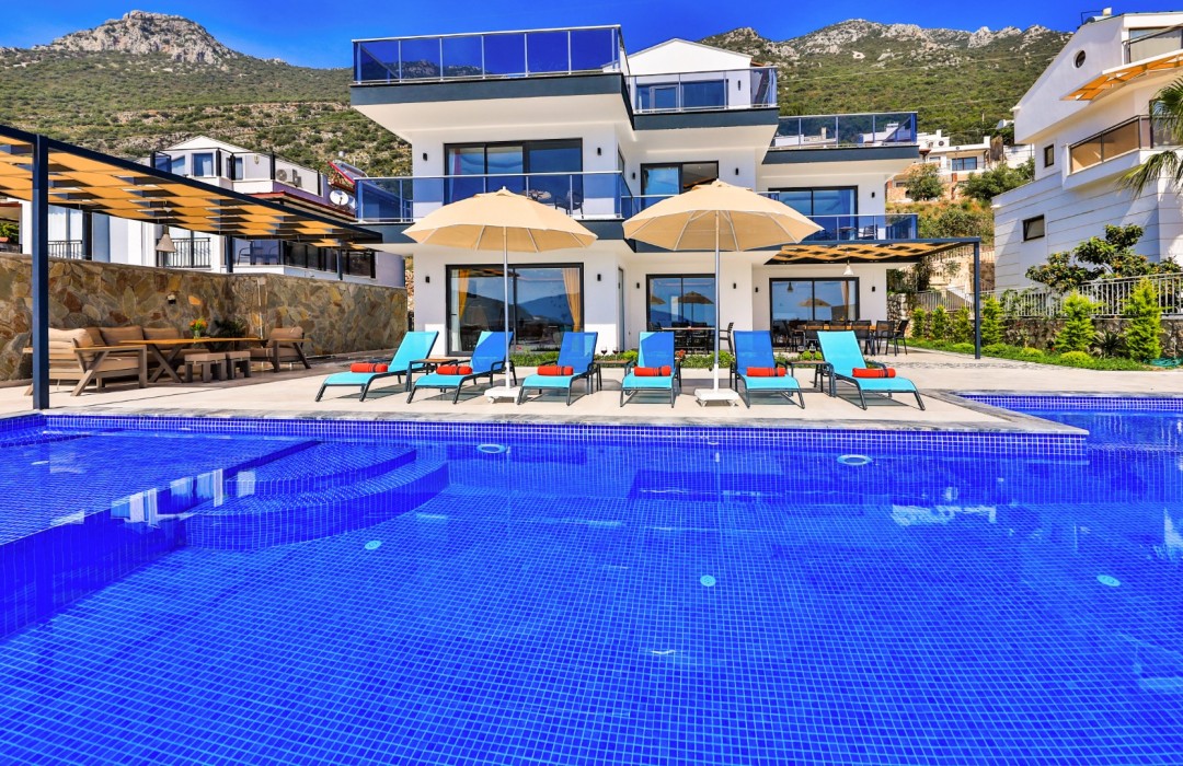 Ön planda mavi mozaikli havuz, havuz kenarında şezlonglar, şemsiyeler ve arkada beyaz katlı villa.