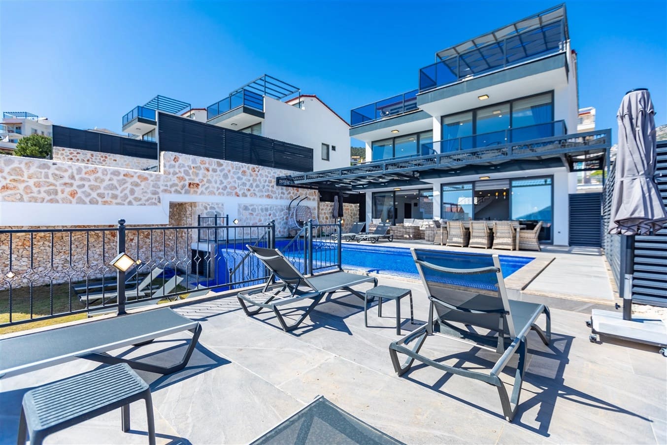 Taş duvarlı terasta şezlonglar ve havuz kenarında modern villa.