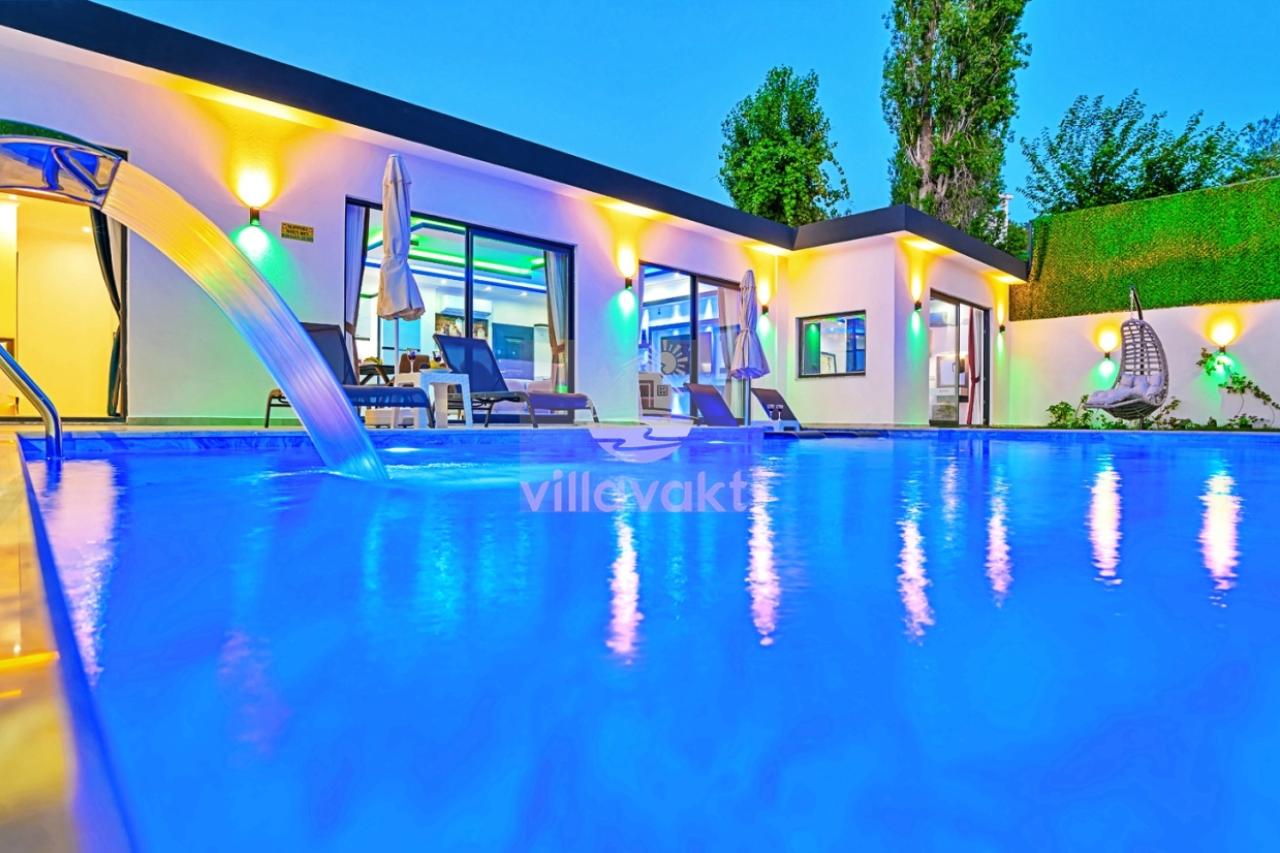 Akşam vakti aydınlatmalı havuz kenarında şezlonglar ve villa dış cephesi.