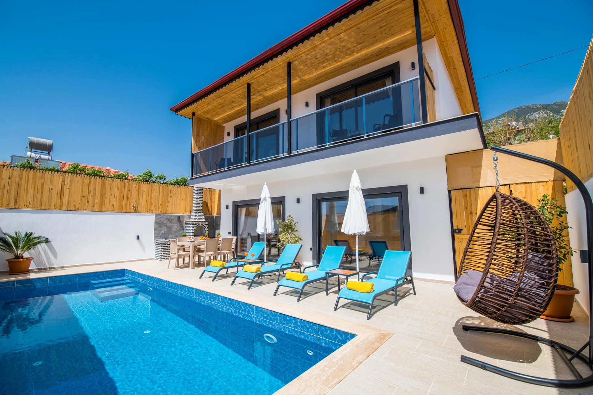Gündüz vakti havuz başında şezlonglar, salıncak ve iki katlı villa cephesi.