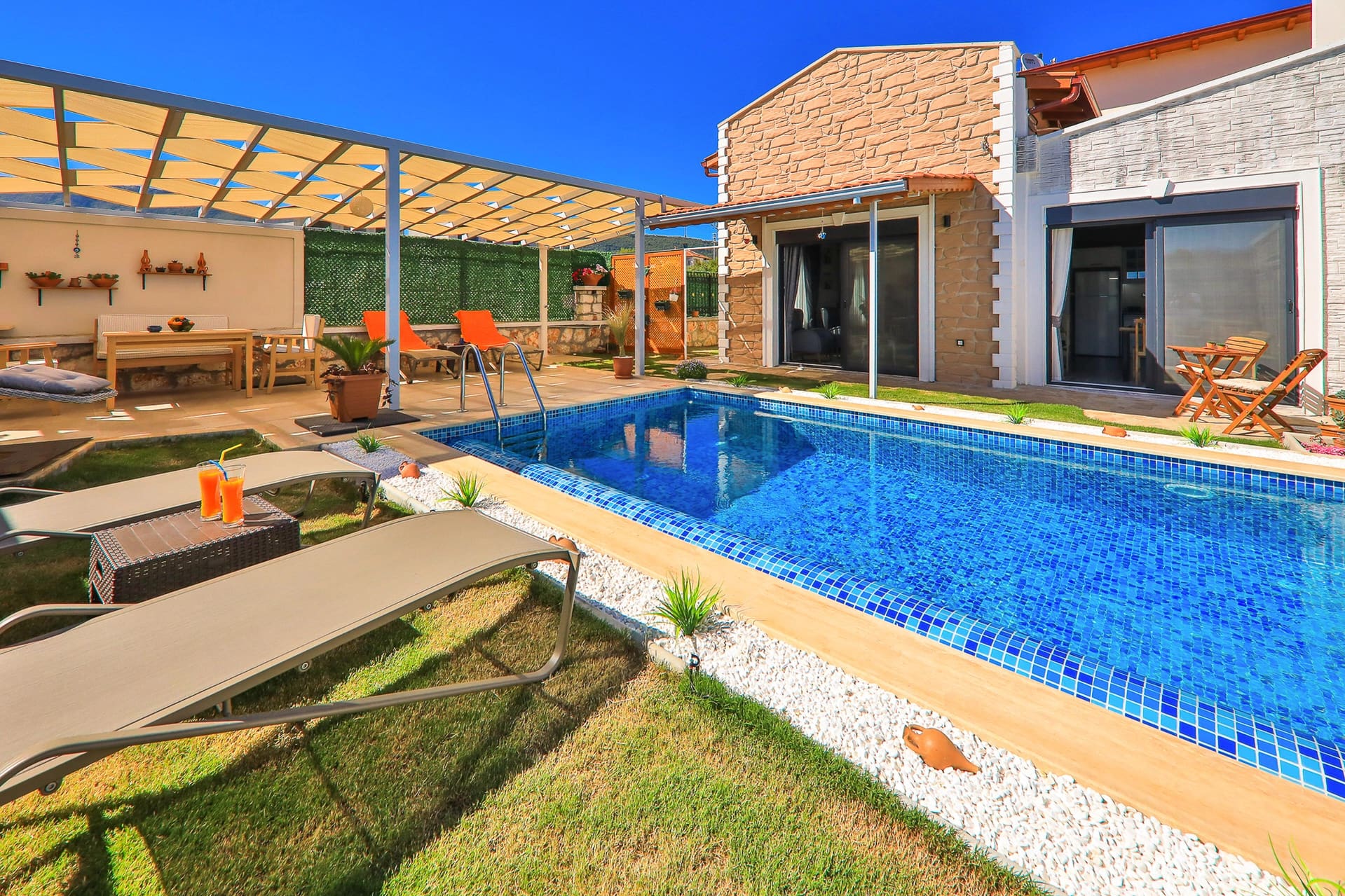 Güneşli havada villa bahçesi, havuz, şezlonglar ve ahşap pergola altında oturma alanı.