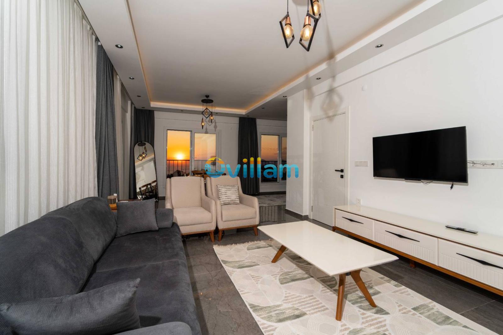 Gün batımı manzaralı pencereleri olan, gri koltuklu ve TV'li oturma odası.