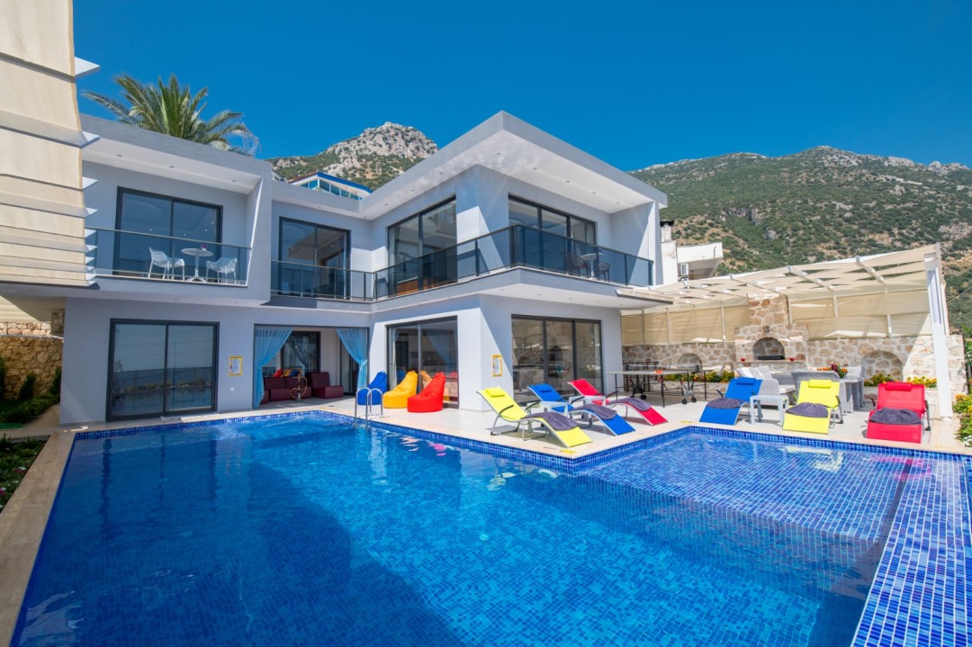 Dağ manzaralı, iki katlı modern villanın önünde mavi fayanslı bir yüzme havuzu ve renkli şezlonglar var.