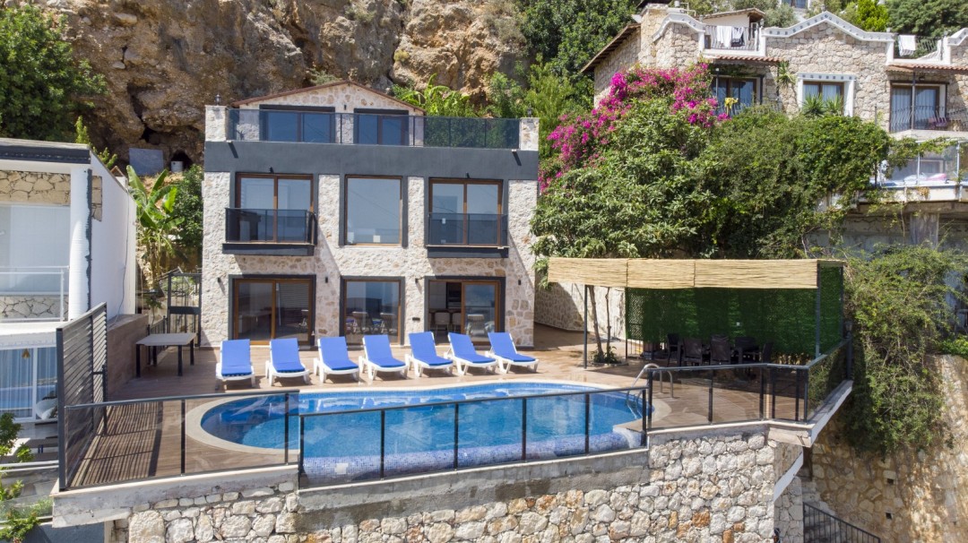 Havuzlu terasında mavi şezlonglar ve gölgelikli yemek alanı bulunan taş cepheli villa.