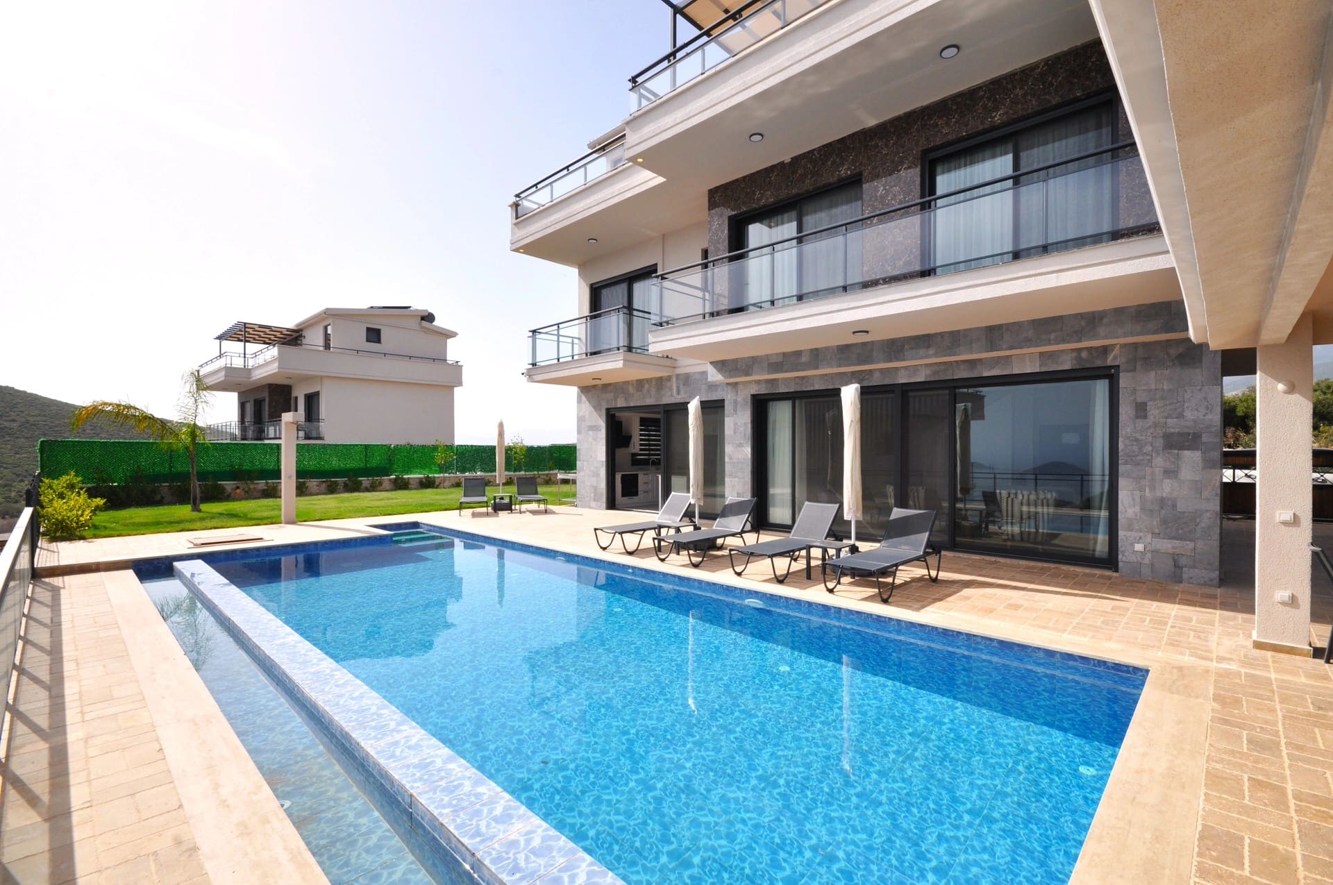Geniş yüzme havuzu ve şezlongları olan iki katlı taş villa dış cephesi.