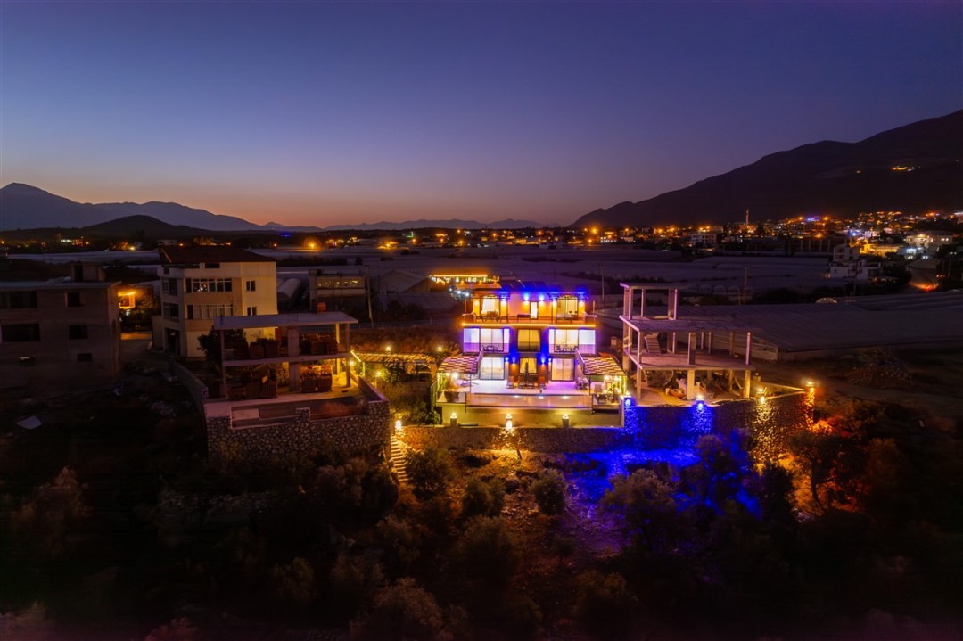 Gece vakti ışıklandırılmış, havuzlu ve teraslı villanın havadan görünümü.