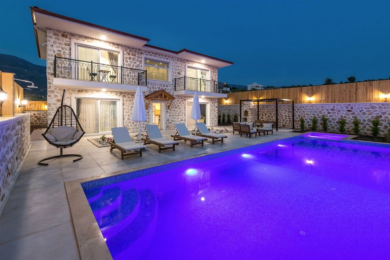 Akşam aydınlatmalı havuz ve taş duvarlı iki katlı villa.