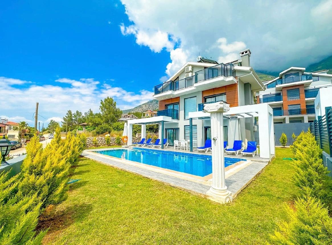 Önünde havuzu ve şezlongları olan çok katlı villa ve yeşil bahçe.
