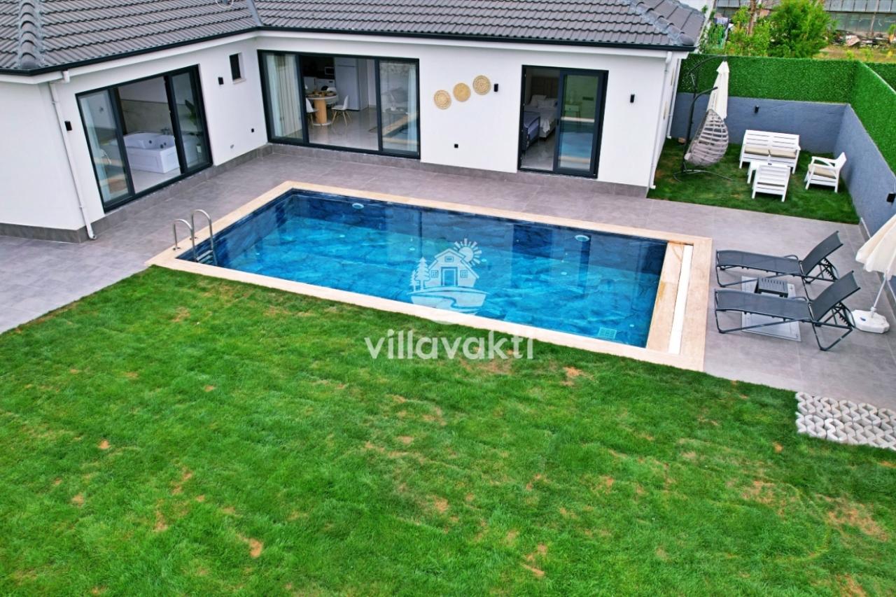 Çim bahçesi, havuzu ve şezlongları bulunan beyaz dış cepheli tek katlı villa.