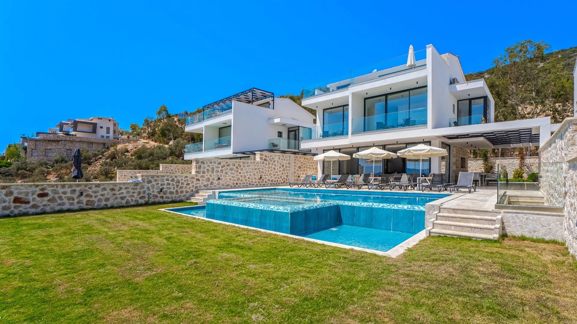 Çim alan ve şezlonglarla çevrili havuzun arkasında yer alan iki katlı beyaz villa.