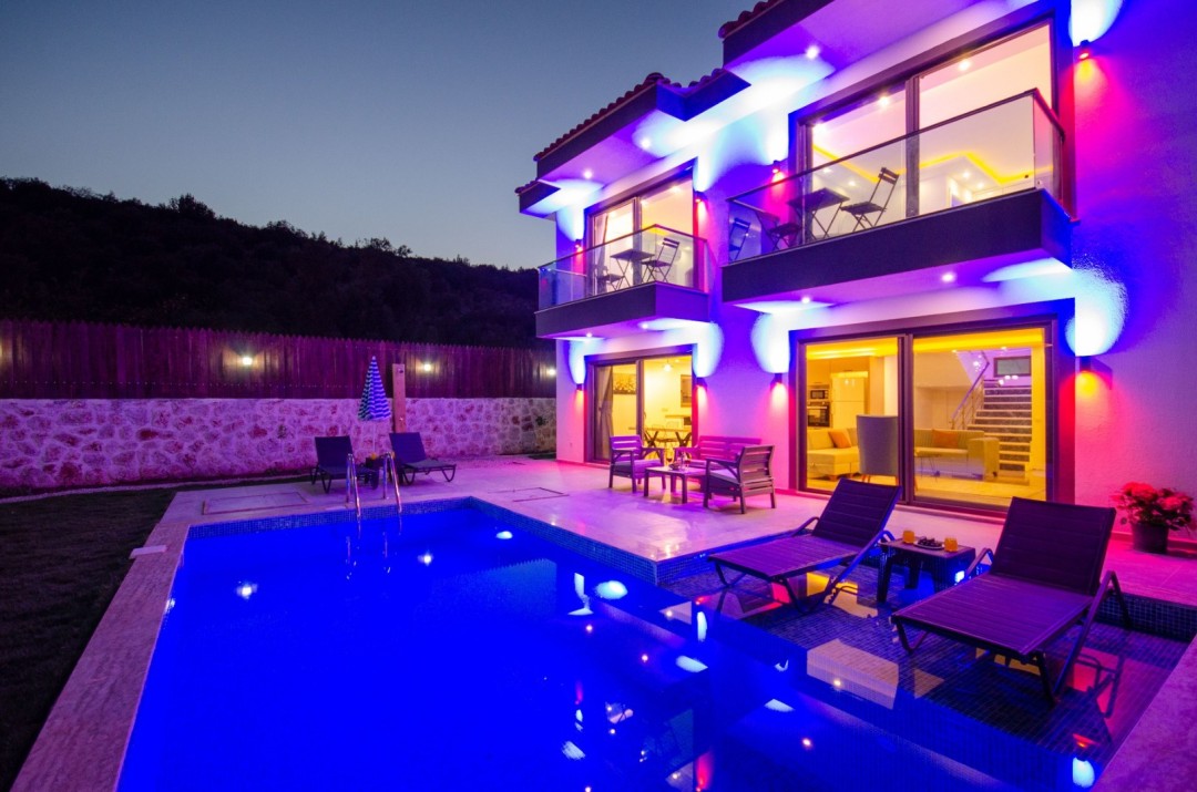 Akşam vakti mor ve mavi ışıklarla aydınlatılmış, havuz başında şezlonglar bulunan iki katlı modern villa.