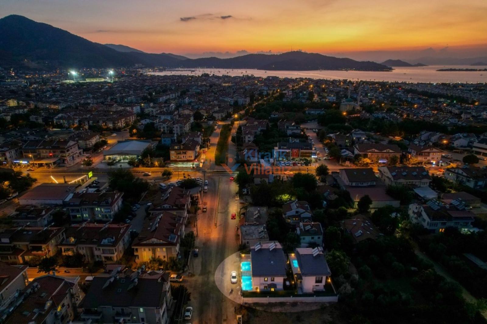 Gün batımında şehir silüeti ve deniz manzarasına bakan villaların havadan görünümü.